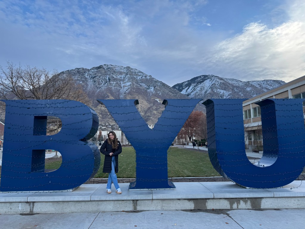 Iris standing outdoors in front of a large blue HOPE sculpture with snowy mountains behind her.
